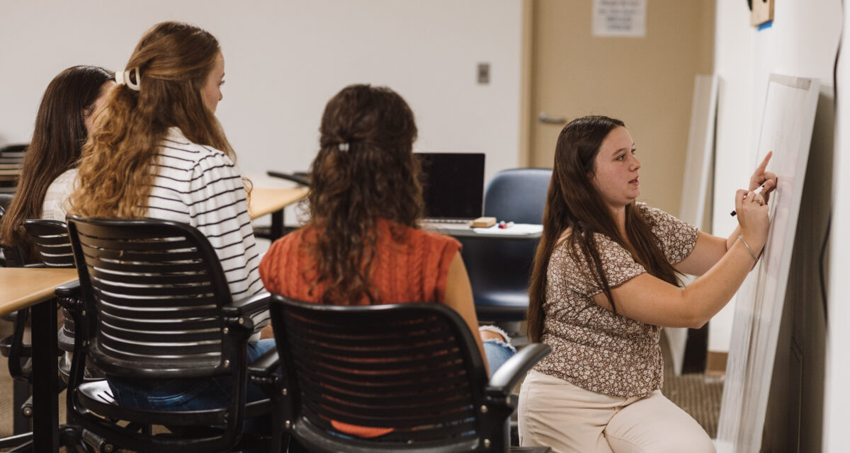 A tutor kneels in front of a whiteboard to help seated students struggling with literacy.