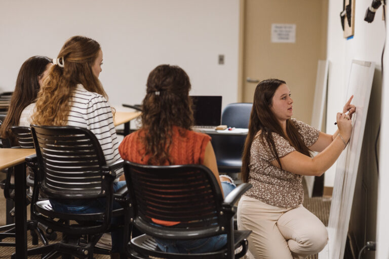 A tutor kneels in front of a whiteboard to help seated students struggling with literacy.